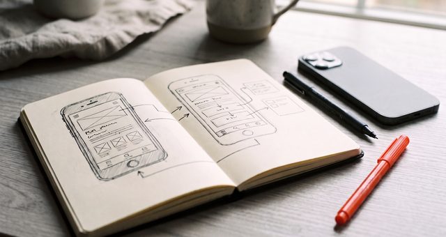 Ein Skizzenbuch mit gezeichneten App-Bildschirmen neben einem Smartphone