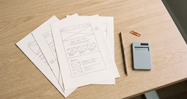 Ausgedruckte Website-Entwürfe, ein Bleistift und ein Taschenrechner auf einem hellen Holztisch